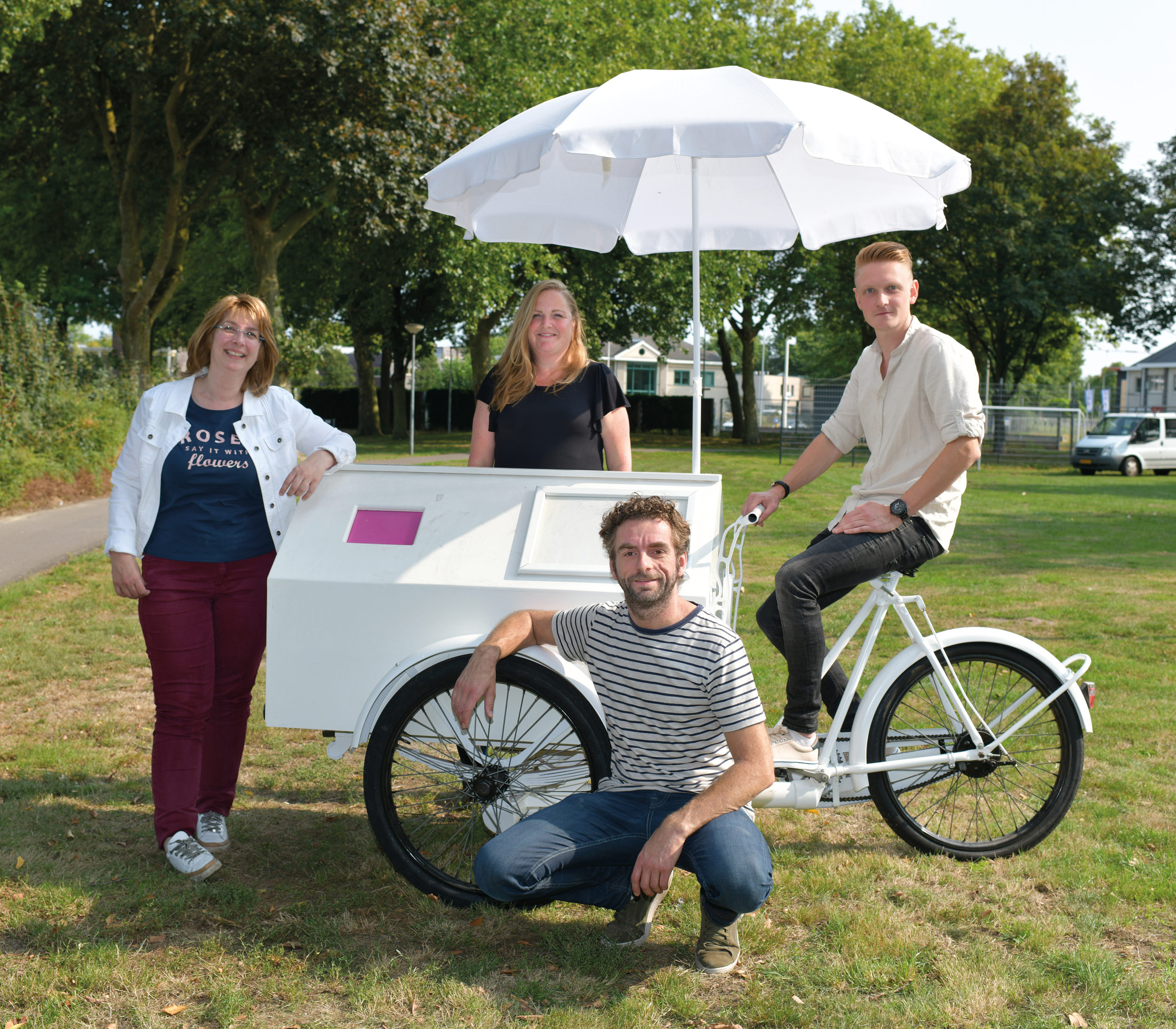 Vier mensen bij een witte bakfiets met parasol in een groene woonomgeving.
